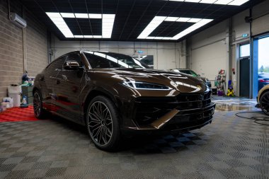 Kahverengi bir Lamborghini Urus. Kapalı bir garajda fayanslı zemini, tuğla duvarları ve parlak floresan ışıkları olan bir garajda. Temizlik malzemeleri görünür..