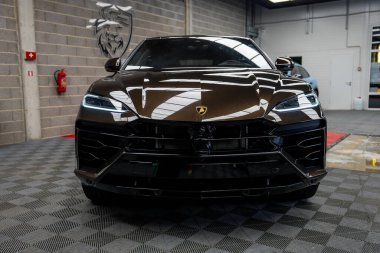 Koyu kahverengi bir Lamborghini Urus beton duvarlar, yangın güvenlik ekipmanları ve arka planda aslan amblemi olan kareli bir zeminde park etmiş..