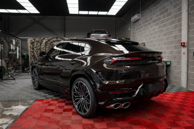 Parlak bir kapağı olan siyah Lamborghini Urus kırmızı kareli bir paspasın üzerine park edilmiştir. Arka planda yükseltilmiş bir platformda ikinci bir araba görülebilir..
