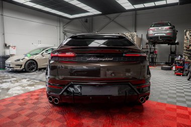 Kırmızı kareli zemine park etmiş siyah Lamborghini Urus, dört kaplamalı egzoz boruları sergiliyor. Bej rengi Porsche Macan ve hidrolik asansördeki araba arka planda görünüyor..