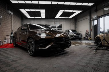 Koyu kahverengi bir Lamborghini Urus modern bir garajda kareli zemin, endüstriyel duvarlar, cam kapılar ve arka planda iki lüks araba ile sergileniyor..