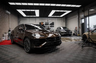 Kahverengi bir Lamborghini Urus modern bir garajda siyah bir Porsche Macan ve bej renkli Bentley Bentayga ile birlikte kareli zeminde sergileniyordu..