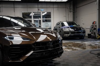 Ön planda kahverengi Lamborghini Urus ve arka planda siyah Porsche Macan olan lüks bir araba tesisi..