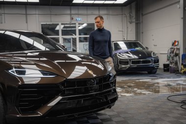 Parlak, siyah bir Lamborghini Urus iyi aydınlatılmış bir garaja park etmiş. Yanında Porsche Macan 'ı olan ve arka planda temizlik ekipmanları olan bir adam var..