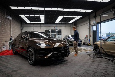 Koyu kahverengi bir Lamborghini Urus kareli zeminli, cam kapılı ve yakındaki aracı kontrol eden bir adamla iyi aydınlatılmış bir galeride sergileniyor..