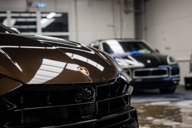 Modern kapalı bir garajda Lamborghini Urus kaputu ve logosunun yakın görüntüsü. Parlak ışık altında bir Porsche Macan görünür..