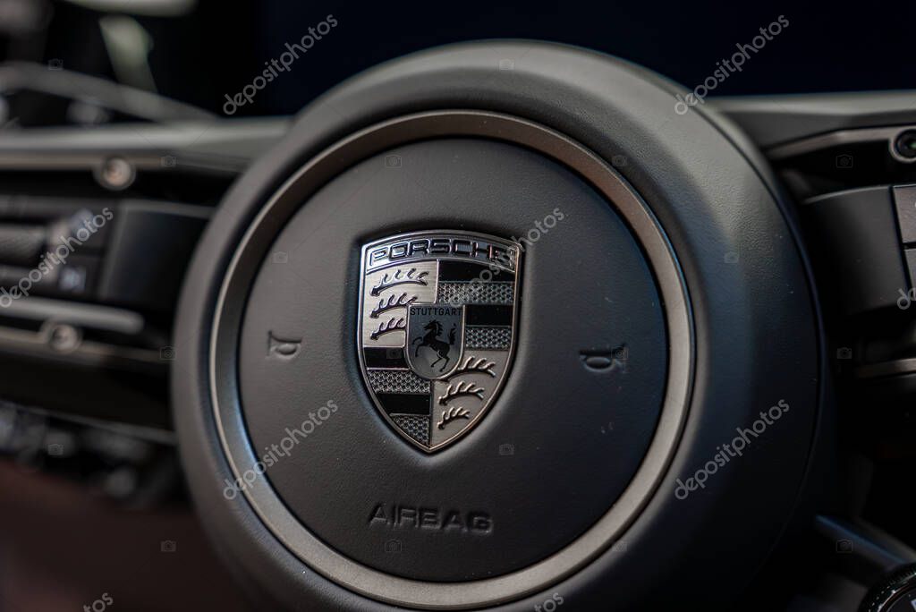 Detailed view of a Porsche steering wheel featuring the Stuttgart crest, sleek dark leather finish, airbag marking, and faint dashboard controls.
