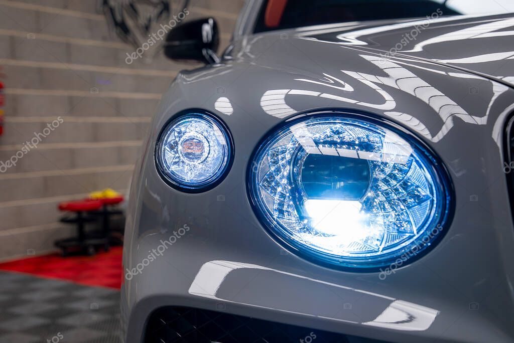 Front headlight assembly of a Bentley Bentayga with illuminated blue LED lights, gray exterior, tiled flooring, and a brick wall in a showroom.