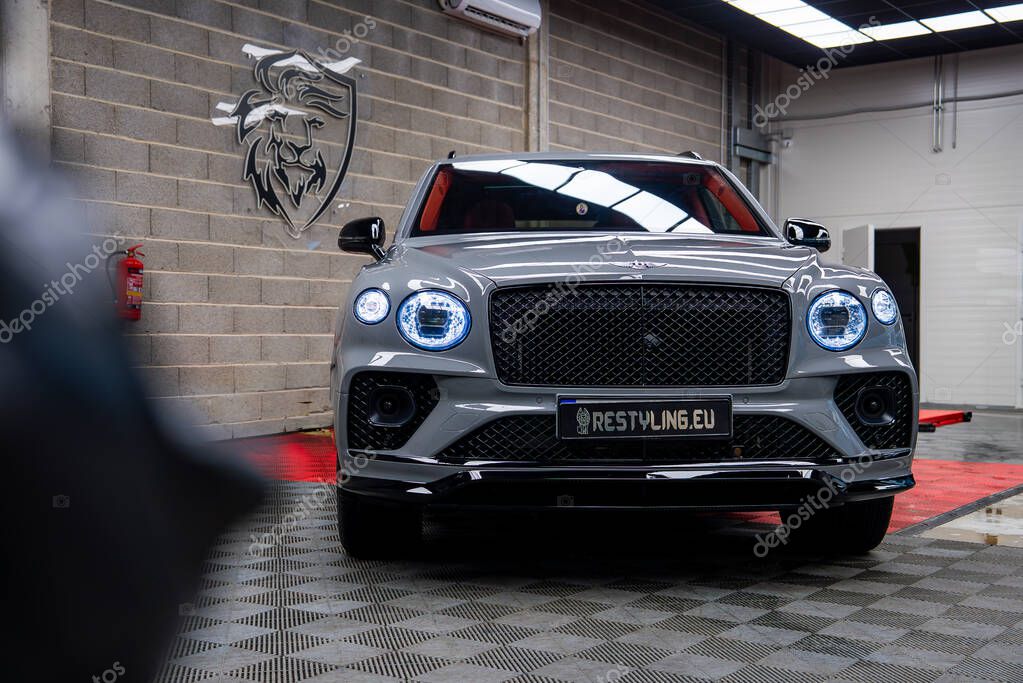 A silver Bentley Bentayga luxury SUV on a checkered floor in a well lit indoor garage. A red platform and lion emblem on a brick wall are visible.