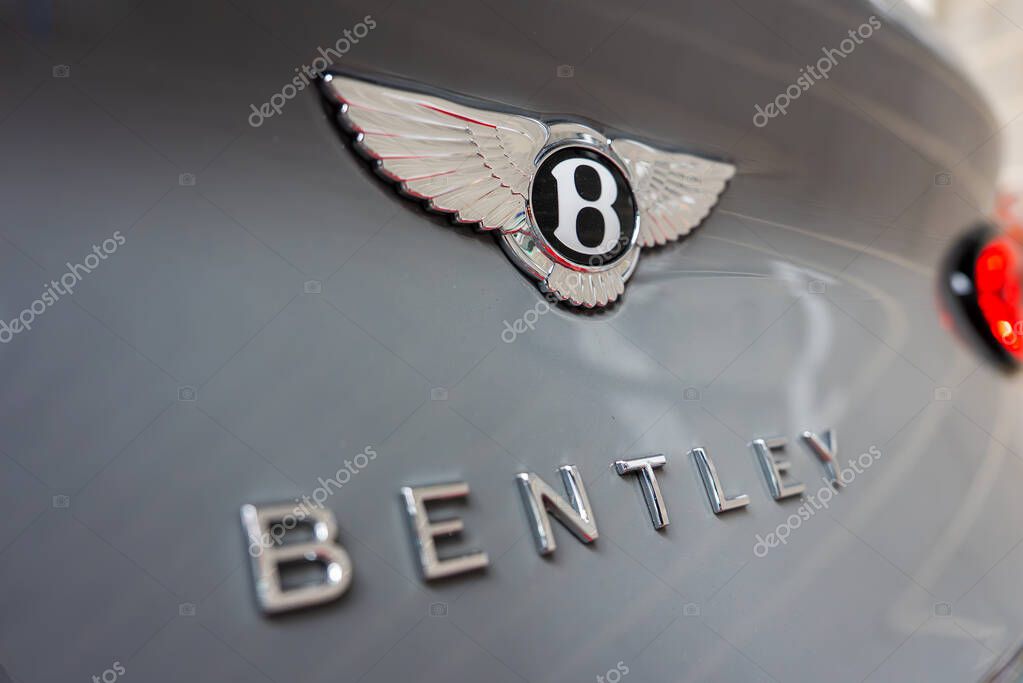 Close up view of the Bentley logo with outstretched wings and polished chrome lettering on a sleek gray surface. Blurred background with red taillight visible.