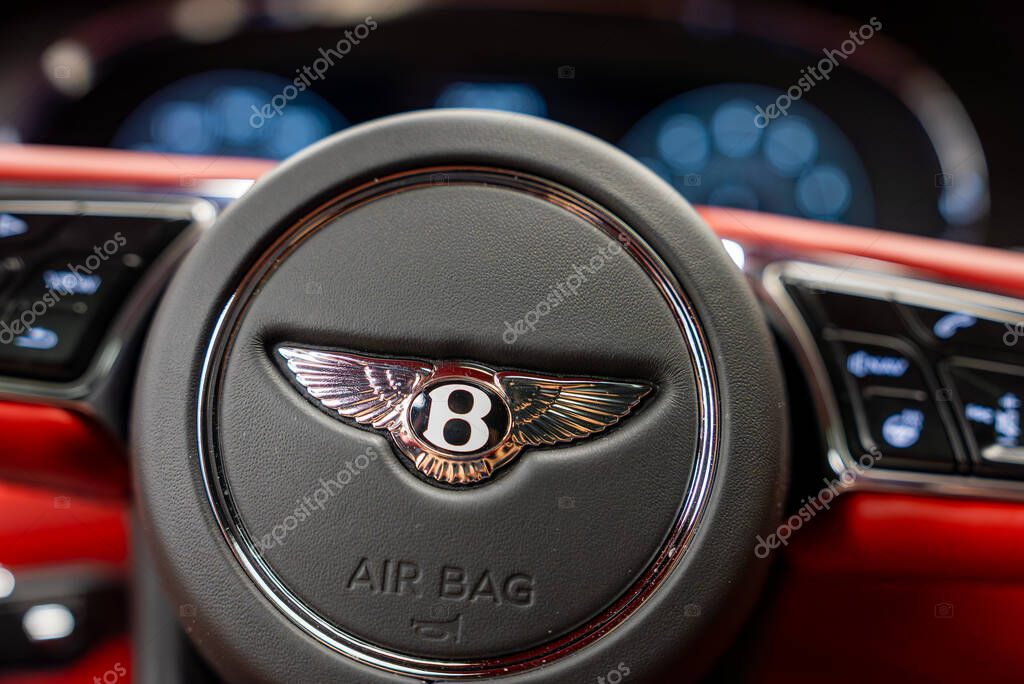 Close up view of a Bentley steering wheel featuring the iconic B logo, surrounded by a red interior, control buttons, and a blurred digital dashboard.