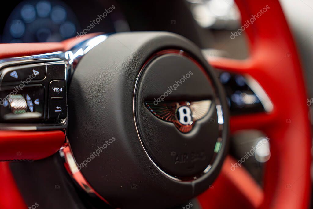 Detailed view of a Bentley steering wheel featuring the iconic B logo, red leather accents, and chrome detailing, with a softly blurred background.