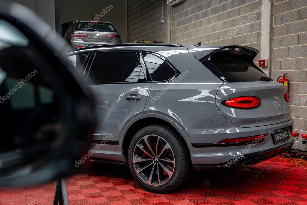 Gray Bentley Bentayga parked on red checkered floor indoors, showcasing rear lights and logo. Background includes car on lift and concrete block walls.