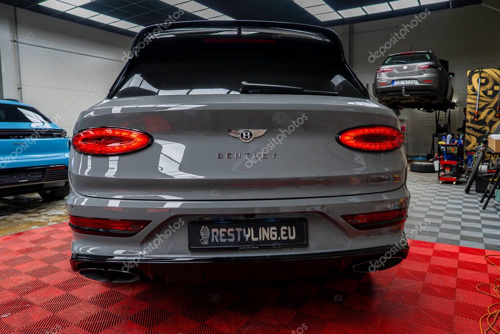Gray Bentley Bentayga parked indoors on red checkered mat with visible Bentley logo. Blue Porsche Macan and car lift visible in clean workshop space.