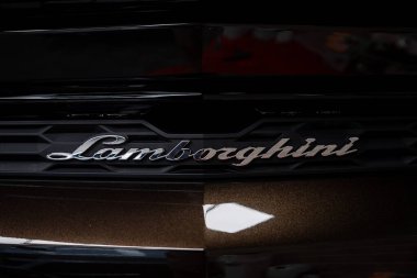 Siyah ızgarada metalik amblemi olan bir Lamborghini 'nin arka kısmı, parlak kahverengi kaplama ve kırmızı beyaz tonlu bulanık sergi odası arka planı..