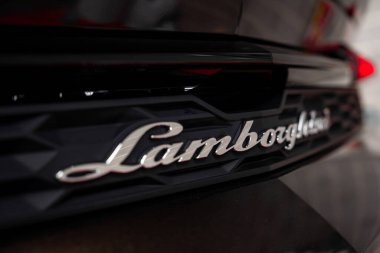 Lamborghini 'nin arka tarafının yakın görüntüsü, parlak siyah bir yüzey üzerindeki gümüş logoyu desenli tasarımla sergiliyor. Arka plan kapalı alanda bulanık görünüyor..