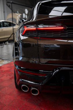 Arka planda siyah Lamborghini Urus çift egzoz boruları ve kırmızı stop lambaları var. Arka planda başka bir lüks araba olan kırmızı bir kata park edilmiş..