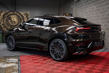 Koyu kahverengi Lamborghini Urus. Kapalı bir yerde, kırmızı ve siyah kareli zeminde parlak bir kapağı var. Arka planda beton duvar ve sanatsal duvar var..