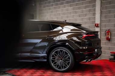 Parlak, koyu kahverengi bir Lamborghini Urus kırmızı kareli zemine park etmiş. Parlak tasarım ve alaşımlı tekerlekler gri beton bir duvara karşı duruyor..