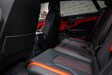 Lamborghini Urus 'un içinde kırmızı aksanlı siyah deri koltuklar, karmaşık dikişler, gelişmiş kontroller ve arka planda beton bir duvar bulunur..