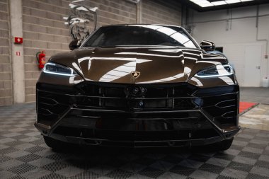 Parlak, koyu kahverengi bir Lamborghini Urus kareli zemine park etmiş. Galeride tuğla duvarlar, yangın söndürücü ve boğa logosu var..