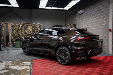 Parlak, siyah bir Lamborghini Urus kırmızı ve gri kareli bir zemine park etmiş. Modern garajda beton duvarlar ve dekoratif duvar panelleri yer alıyor..