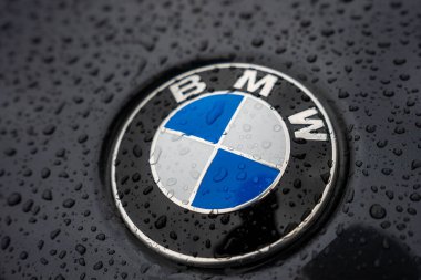 Islak bir araç kaputunda BMW logosu, su damlacıkları ve parlak yüzeye yansıyan soluk bir siluet..
