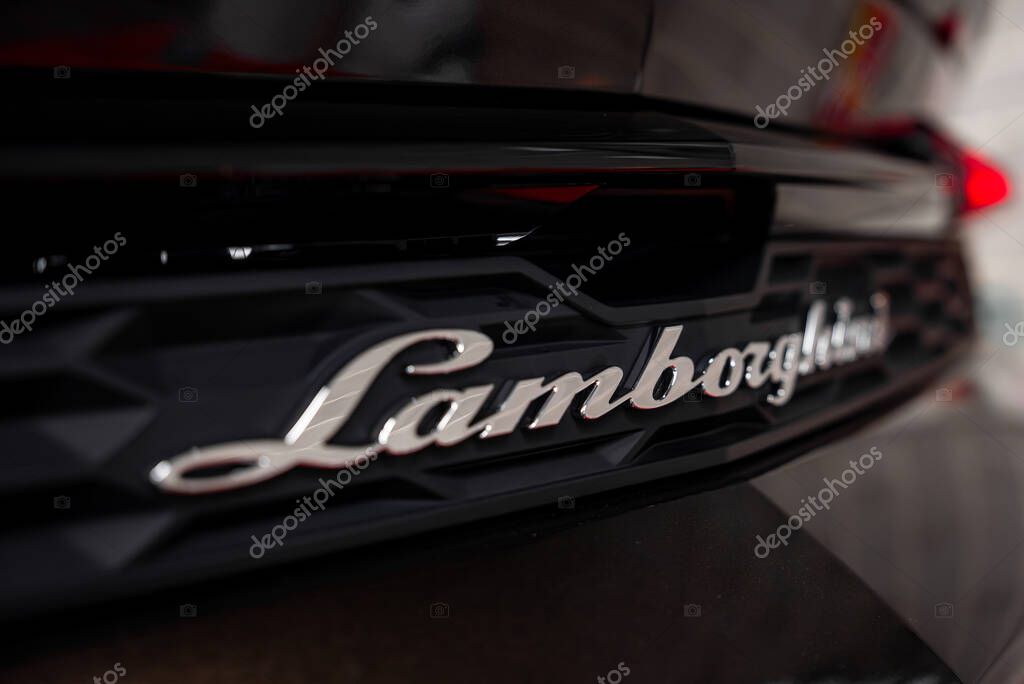 Close up view of a Lamborghini rear, showcasing the silver logo on a glossy black surface with textured design. Background appears blurred indoors.