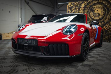 Kırmızı bir 2025 Porsche 911 GT3922 kaputta koruyucu ambalaj, siyah tekerlekler ve mavi ayna aksanlı, atölyede altın bir duvar resmine karşı..
