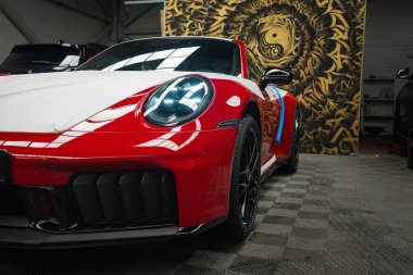 Kırmızı bir 2025 Porsche 911 GT3922 'nin ön ucu LED farları ve aerodinamik ızgarası olan, kareli bir zemine park edilmiş,.