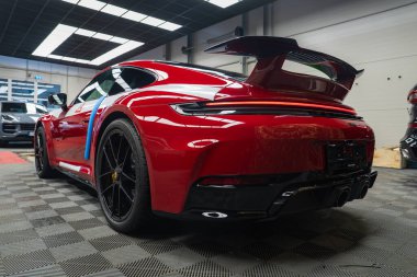 Kırmızı bir 2025 Porsche 911 GT3922 arka kanadı, siyah alaşımlı tekerlekleri ve çift egzozlu kareli zemini ve şebeke aydınlatması olan bir galeride duruyor..