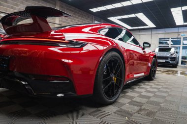 Kırmızı bir 2025 Porsche 911 GT3922 modern bir sergi salonunda sergilenmektedir. Özellikler arasında bir arka kanat, siyah alaşımlı tekerlekler ve kareli zemin tasarımı yer alıyor..