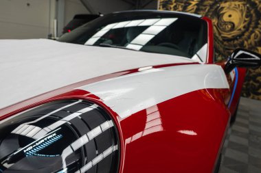 Kırmızı bir 2025 Porsche 911 GT3922 'nin ön bölümü. Kapalı mekanda parlak farlar, HD Matrix metni, beyaz bir koruyucu ambalaj ve altın bir duvar resmi sergileniyor..