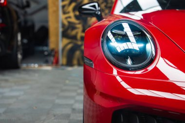 Kırmızı bir 2025 Porsche 911 GT3922 'nin ön tarafı, parlak LED farları aydınlatıyor. Görünür ekipmanlarla bulanık garaj arka planı.