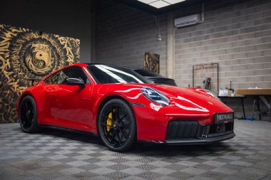 Canlı kırmızı bir 2025 Porsche 911 GT3922 kapalı bir yere park edilmiş siyah tekerlekler, sarı fren kalibreleri ve endüstriyel stil dekor..