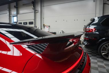 Kırmızı bir 2025 Porsche 911 GT3922 'nin arka kanadı siyah arka kanadı, modern bir sergi salonunda fayanslı döşemeleri ve kısmen görünür siyah bir SUV' u var..