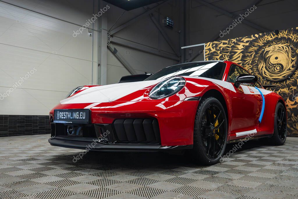 A red and white 2025 Porsche 911 GT3 922 with black wheels and yellow brake calipers displayed indoors on a checkered floor near a decorative wall panel.