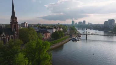 Frankfurt, Almanya 'nın havadan görünüşü, Ana Nehir' i, modern gökdelenleri, Eiserner Steg 'i, rıhtıma yanaşmış tekneleri ve kırmızı çan kulesi olan bir kiliseyi gösteriyor..