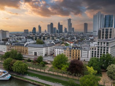 Frankfurt, Almanya 'nın havadan görünüşü, Commerzbank Kulesi gibi modern gökdelenler, bir nehir kenarı gezinti güvertesi, rıhtımlı bir tekne ve dramatik bir günbatımı gökyüzü..