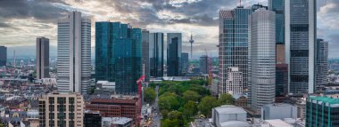 Frankfurt, Almanya 'nın havadan görünüşü, Ana Kule, Europaturm, modern gökdelenler, yeşil bir park, ve güneş ışığıyla aydınlanan bulutlu bir gökyüzü.