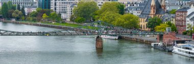 Eiserner Steg yaya köprüsü, Almanya 'nın Frankfurt kentindeki Main River boyunca uzanır. Arka planda ikiz kuleleri olan bir kilise de dahil olmak üzere çağdaş ve tarihi mimari, yeşillik ve nehir kenarındaki gezinti güvertesiyle birlikte görülebilir..