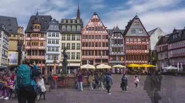 Frankfurt, Almanya 'daki Romerberg Meydanı' nda yarı keresteli binalar, Adalet Çeşmesi ve sarı şemsiyeler altında dinamik yaya faaliyetleri yer alıyor..
