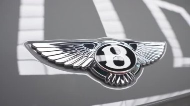 Bentley logosunun yansıtıcı kaputunda gümüş kanatlar olan yakın görüntüsü. Işık desenleri gösterişli tasarımı vurguluyor, lüks bir araba ayarını işaret ediyor..