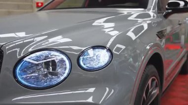 Kamera, kırmızı aksanlı gri Lamborghini Urus boyunca döner, parlak kaplamasını ve aerodinamik tasarımını iyi aydınlatılmış bir sergi salonunda gösterir..