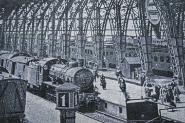 Frankfurt 'taki Hauptbahnhof' da bir buhar lokomotifi. Etrafı yolcular ve işçilerle çevrili. Kemerli çelik çatı ve klasik tren tasarımı dönemin altını çiziyor..
