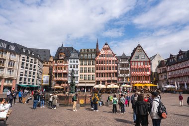 Frankfurt 'taki Romerberg Meydanı' nda geleneksel yarı ahşap binalar, Adalet Çeşmesi, kaldırım taşı zemin ve şemsiyelerin altında açık hava koltukları yer alıyor..