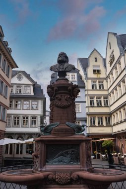 Friedrich Schiller heykeli, Frankfurt 'ta parçalı bulutlu bir gökyüzü altında Avrupa tarzı binalarla çevrili süslü bir çeşmenin tepesinde..