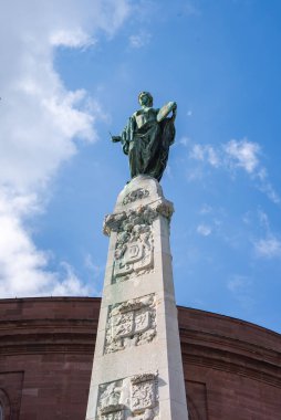 Frankfurt, Almanya 'da bulutlu mavi bir gökyüzüne karşı, tablet taşıyan bronz bir heykel ve karmaşık oymalarla dolu uzun bir taş sütun..