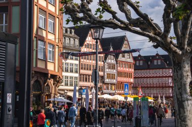 Frankfurt, Almanya 'daki Romerberg Meydanı' nda, güneşli bir günde kırmızı ve beyaz yarı kereste binalar, canlı bir kalabalık, çıplak ağaçlar, sokak tabelaları ve sokak lambaları yer alıyor..