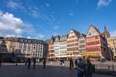 Frankfurt 'taki Romerberg Meydanı' nda renkli yarı ahşap binalar, Frankfurt Katedrali kulesi ve canlı açık alanların keyfini çıkaran insanlar yer alıyor..